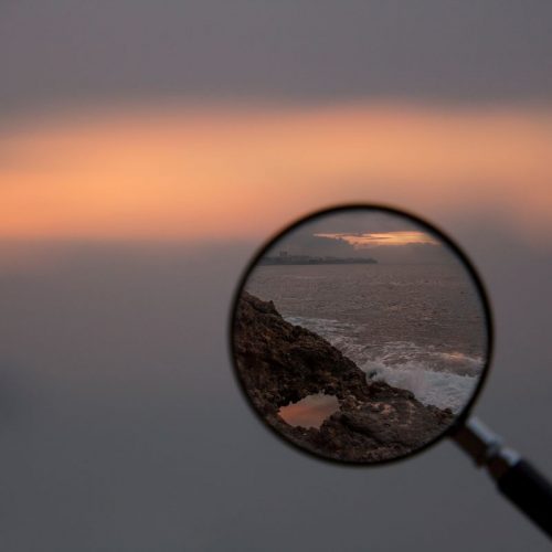 Faith+Lead | A blurred photo of a scene with a magnifying glass providing clarity; through it, a rocky coastline with waves crashing against it is visible.